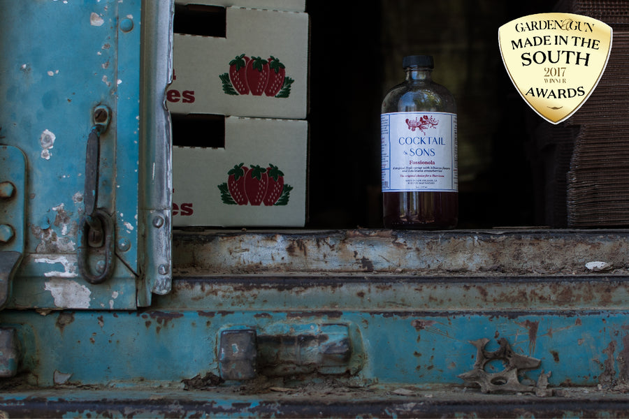 Cocktail & Sons Award Winning Fassionola Cocktail Syrup sitting in the bed of an old truck next to a couple flats of Louisiana Strawberries